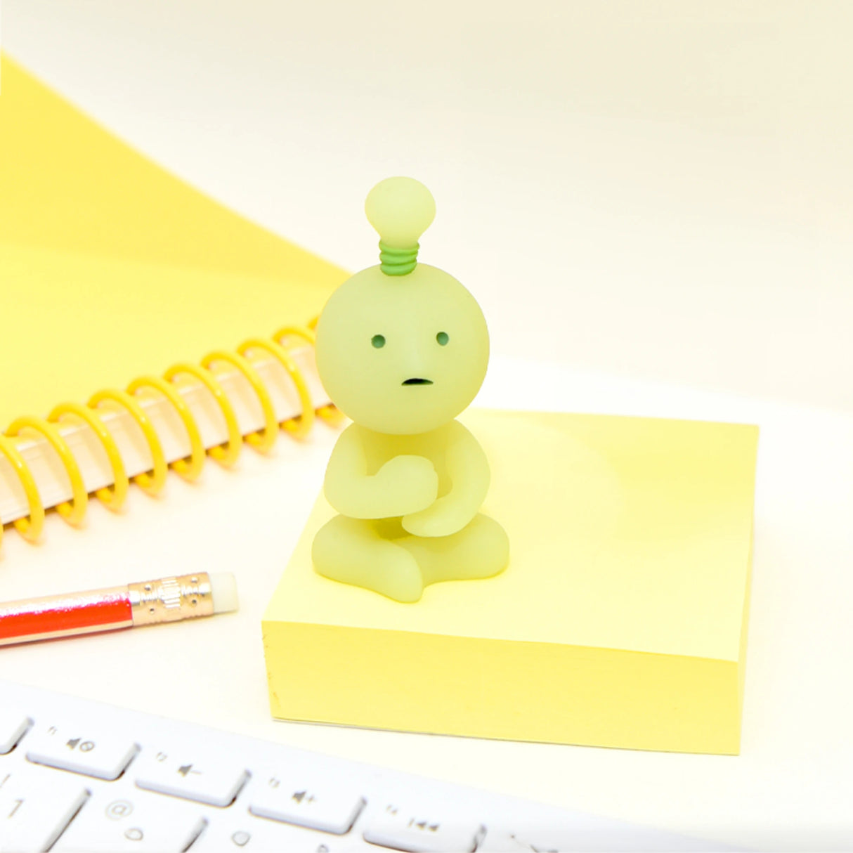 A small, green figurine from the Smiski At Work Series - Blind Box by Smiski sits on a yellow notepad beside a red pencil and a spiral notebook. A white keyboard is partially visible in the foreground, adding to the mystery of this blind box collection.