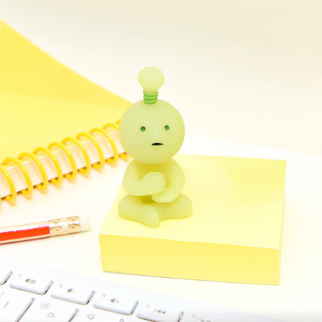 A small, green figurine from the Smiski At Work Series - Blind Box by Smiski sits on a yellow notepad beside a red pencil and a spiral notebook. A white keyboard is partially visible in the foreground, adding to the mystery of this blind box collection.