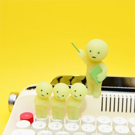 Four small, round, pale yellow figurines from the Smiski At Work Series - Blind Box by Smiski are perched on a typewriter. One of them holds a book and points upward. These charming characters add a touch of whimsy to your workspace and might even glow-in-the-dark when the lights go out.