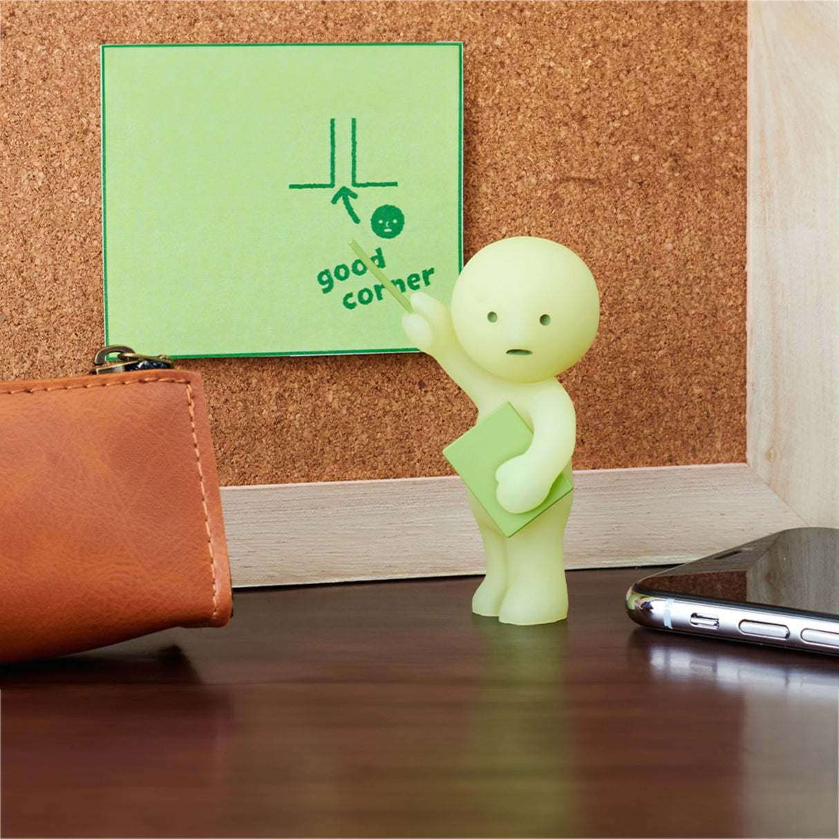 A figurine from the Smiski At Work Series - Blind Box by Smiski is shown holding a green sticky note and gesturing towards another note on a corkboard. Nearby, there's a leather item and a smartphone on the wooden surface, perfectly showcasing the whimsical allure of these popular blind box characters.