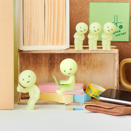 The Smiski At Work Series - Blind Box by Smiski features small green figurines in various office settings with items like a smartphone, wallet, and stationery. One glow-in-the-dark figurine is seated on a stack of erasers with a laptop, while others are positioned nearby in this delightful collection.