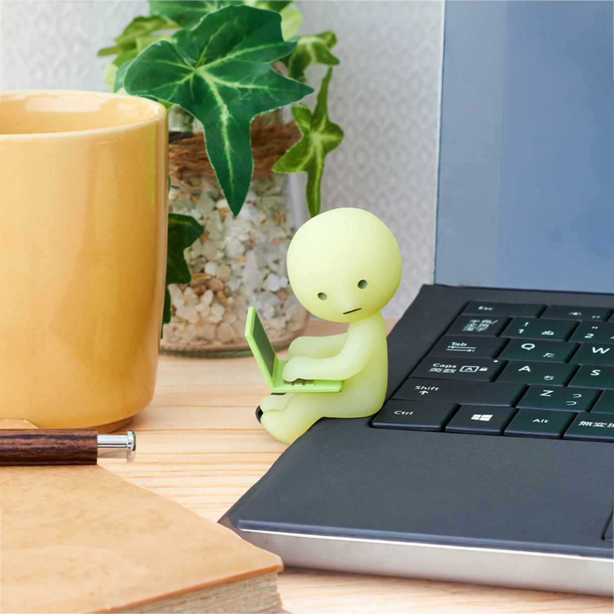 A small glow-in-the-dark figurine from the Smiski At Work Series - Blind Box by Smiski sits on a laptop keyboard, accompanied by a yellow mug, a plant, and a notebook.