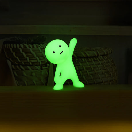 A small, glowing green figure from the Smiski Yoga Series - Blind Box by Smiski stands with one arm raised in a calming yoga pose on a flat surface, while wicker baskets quietly accentuate the background.