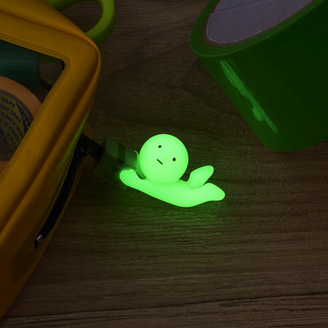 A small, green Smiski ZipperBite - Falling Down figure from Smiski, shaped like a smiling ghost and glowing in the dark, lies on a wooden surface near green tape and a yellow object.