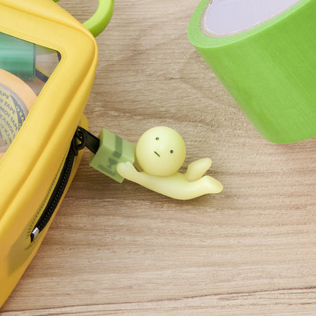 A Smiski ZipperBite - Falling Down keychain from Smiski is attached to the zipper of a yellow pouch next to a roll of green tape on a wooden surface.