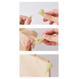 A three-step image highlights the Smiski ZipperBite in the Hugging Knees design, showcasing a vibrant green zipper pull. It attaches seamlessly to a beige bag zipper, making it easy to open. This glow-in-the-dark accessory from Smiski adds a touch of both functionality and fun to your daily essentials.