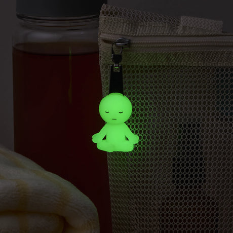 The Smiski ZipperBite - Lotus Pose by Smiski is a small, glow-in-the-dark figure in lotus pose clipped to a beige mesh bag, with a bottle and folded towel nearby, adding peaceful charm to your accessories.