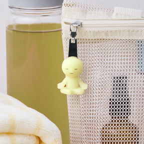 The Smiski ZipperBite - Lotus Pose by Smiski is a small, yellow, glow-in-the-dark figurine attached to a mesh zippered pouch, perfect for adding charm to your accessories. A yellow bottle and folded towel are in the background.