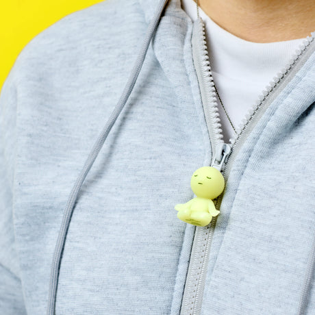 Close-up of a person wearing a grey hoodie with the Smiski ZipperBite - Lotus Pose by Smiski, a small glow-in-the-dark zipper accessory, attached to the zipper against a yellow background.