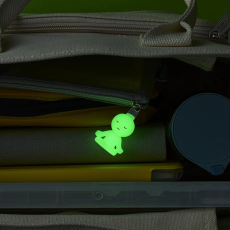The Smiski ZipperBite - Lotus Pose keychain from Smiski glows in the dark as it hangs from a zipper inside a book-filled bag with a round container.