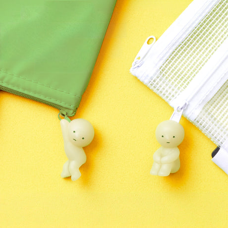 Against a vibrant yellow background, a green pouch and a white mesh pouch feature two small Smiski ZipperBite - Hugging Knees figurines in pale green, hanging from their zippers. These delightful accessories by Smiski bring character with their glow-in-the-dark feature.