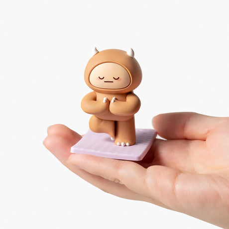 Hand holding a small brown Smoko Hank Yeti figure in a yoga pose on a lavender mat against white background