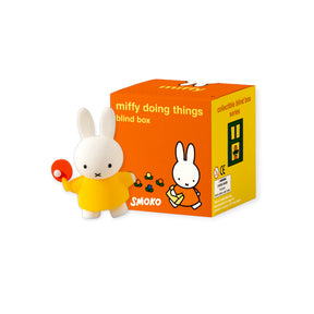 Small white bunny figurine in yellow outfit holding a red ping pong paddle next to orange and yellow blind box