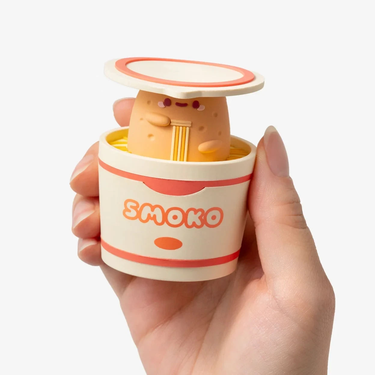 Hand holding a Smoko Tayto Ramen Blind Box vinyl figure partially inside its ramen cup packaging with the lid open.