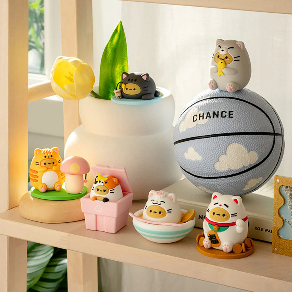 A variety of adorable cat-themed figurines, featuring a Smoko Tayto Potato Cat Blind Box collectible, are displayed on a shelf alongside a small decorative basketball and a vase holding a yellow tulip.