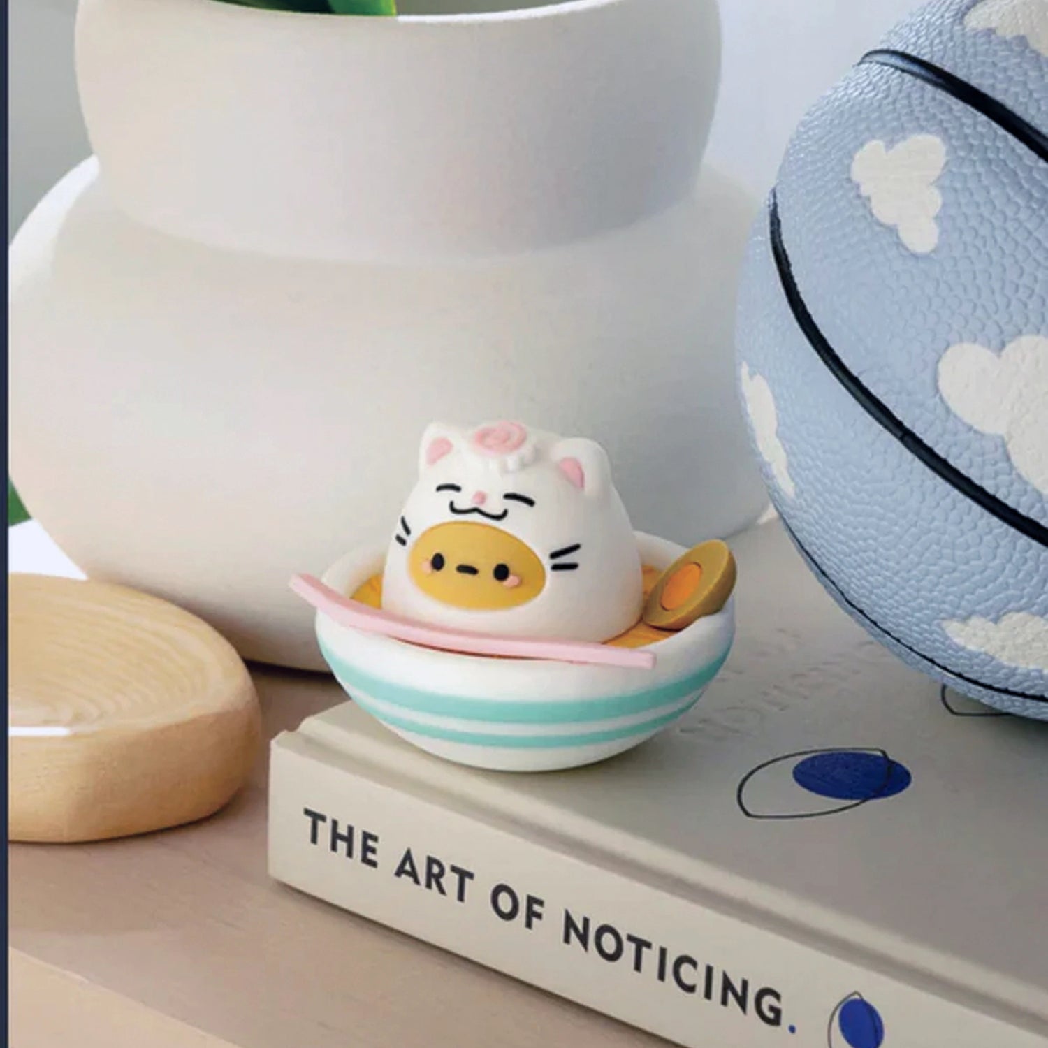 A Smoko Tayto Cat Blind Box figurine in a pig costume sits in a blue and white striped bowl next to a textured basketball, perched on "The Art of Noticing.