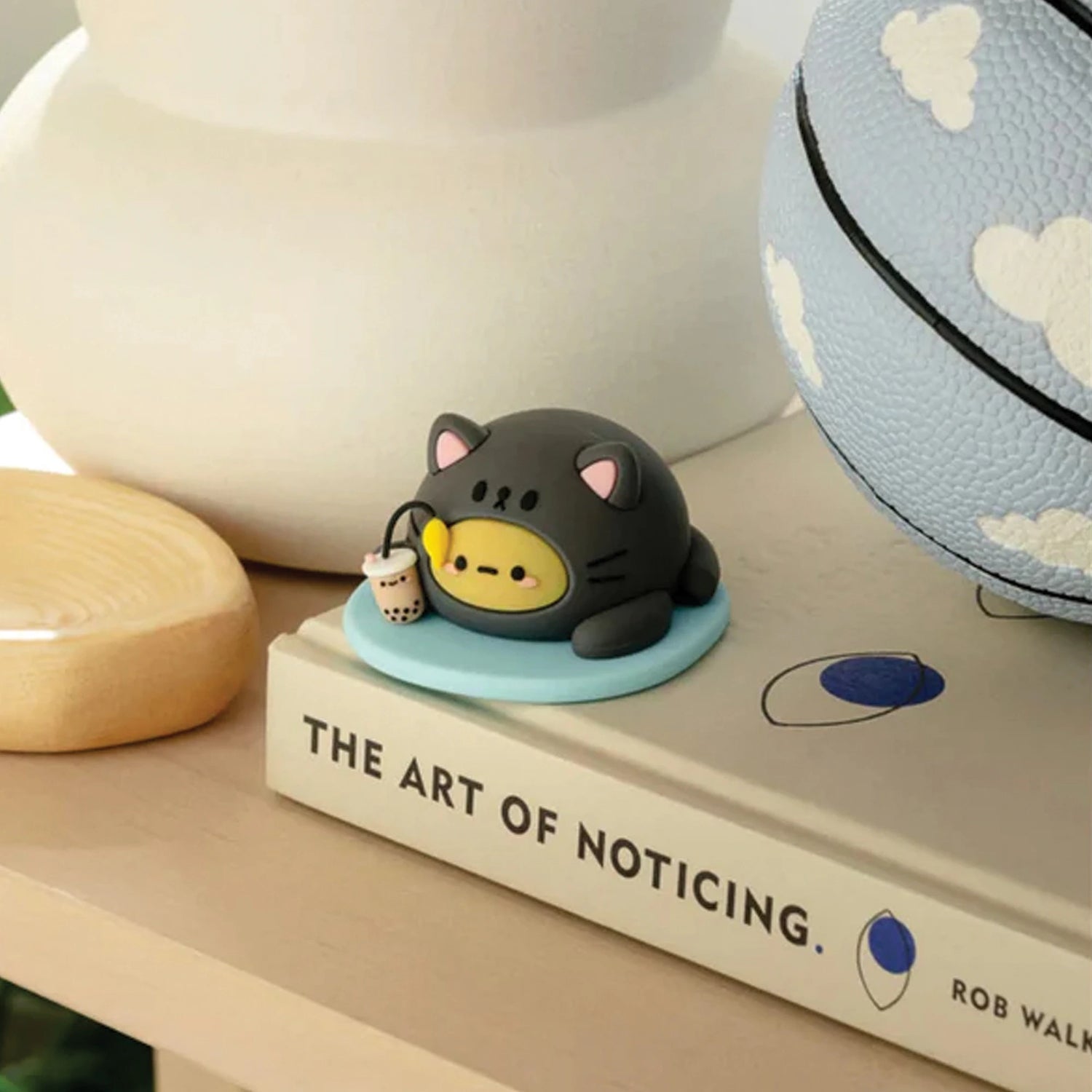 A small, round Smoko Tayto Cat Blind Box figurine holding bubble tea sits on a blue base atop a book titled "The Art of Noticing," with a patterned ball nearby, capturing the playful essence of this Smoko cat-themed Tayto.