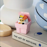 A Smoko Tayto Potato Cat Blind Box collectible figurine and a cow sit in a pink recycling bin on a book titled "The Art of Noticing," next to a basketball with cloud designs.