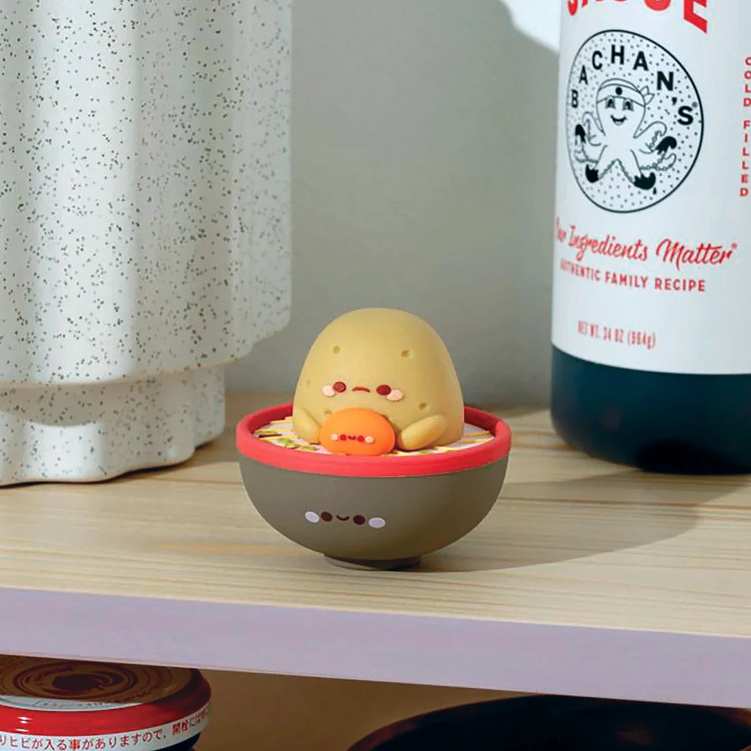 A Smoko Tayto Ramen Blind Box Vinyl Figure rests in a bowl on a wooden shelf, next to a speckled ceramic vase and a bottle with a colorful label, echoing the delightful surprises of every Smoko Blind Box collection.