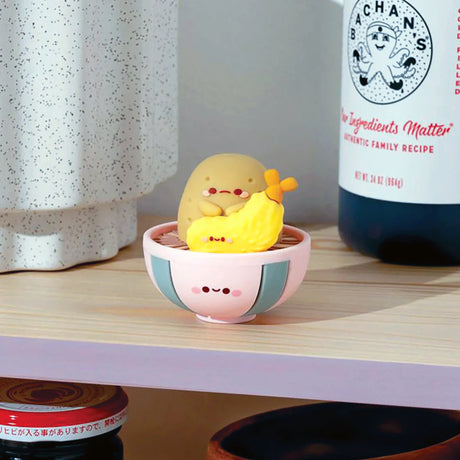 A charming Smoko Tayto Potato Ramen Blind Box figurine, known for its adorable face, rests in a pink bowl beside a smiling banana vinyl figure, showcased on a shelf among other delightful items.