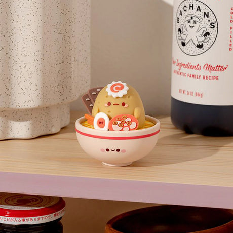 A small Smoko Tayto Potato Ramen Blind Box figure from the Smoko brand resembling food sits in a decorative bowl on a wooden shelf, nestled next to a ceramic container and a bottled product labeled "Bachan's," adding a whimsical touch to the display.