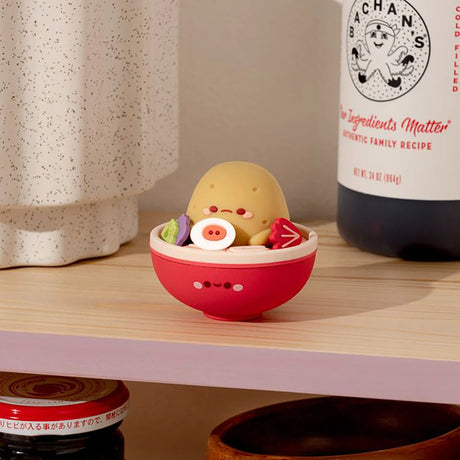 A charming figurine from the Smoko Tayto Potato Ramen Blind Box collection, featuring a potato character styled in a bowl with various food items, is displayed on a shelf next to a speckled container and sauce bottle.