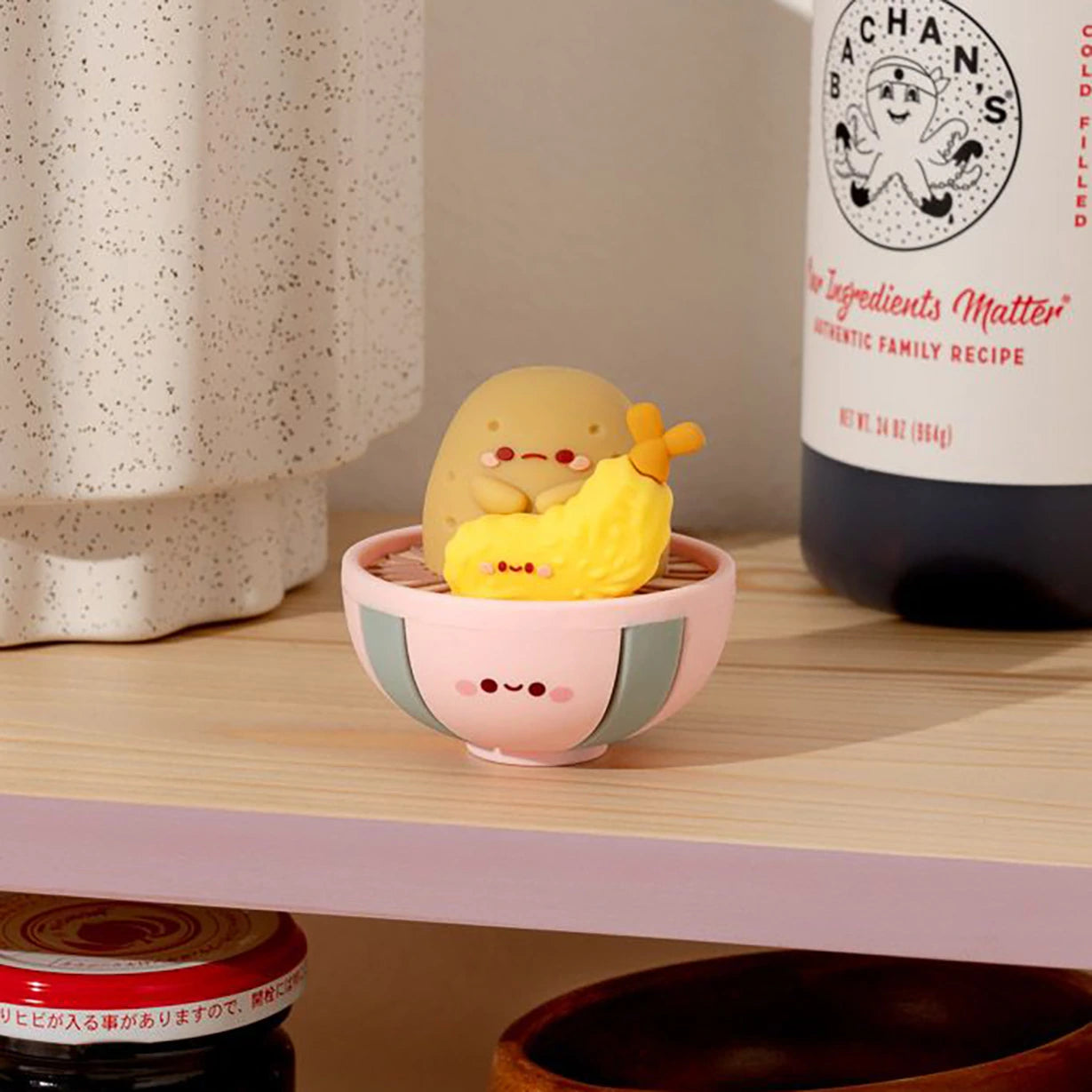 Adorable Smoko figures sit in a pink bowl on a shelf, complemented by a beige jar and sauce bottle in the background. The Smoko Tayto Potato Ramen Blind Box brings an extra dash of charm to the scene.