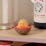 The Smoko Tayto Potato Ramen Blind Box features a petite, round, yellow figure comfortably situated in a ramen-like bowl on a wooden shelf, flanked by a speckled container and a branded bottle.