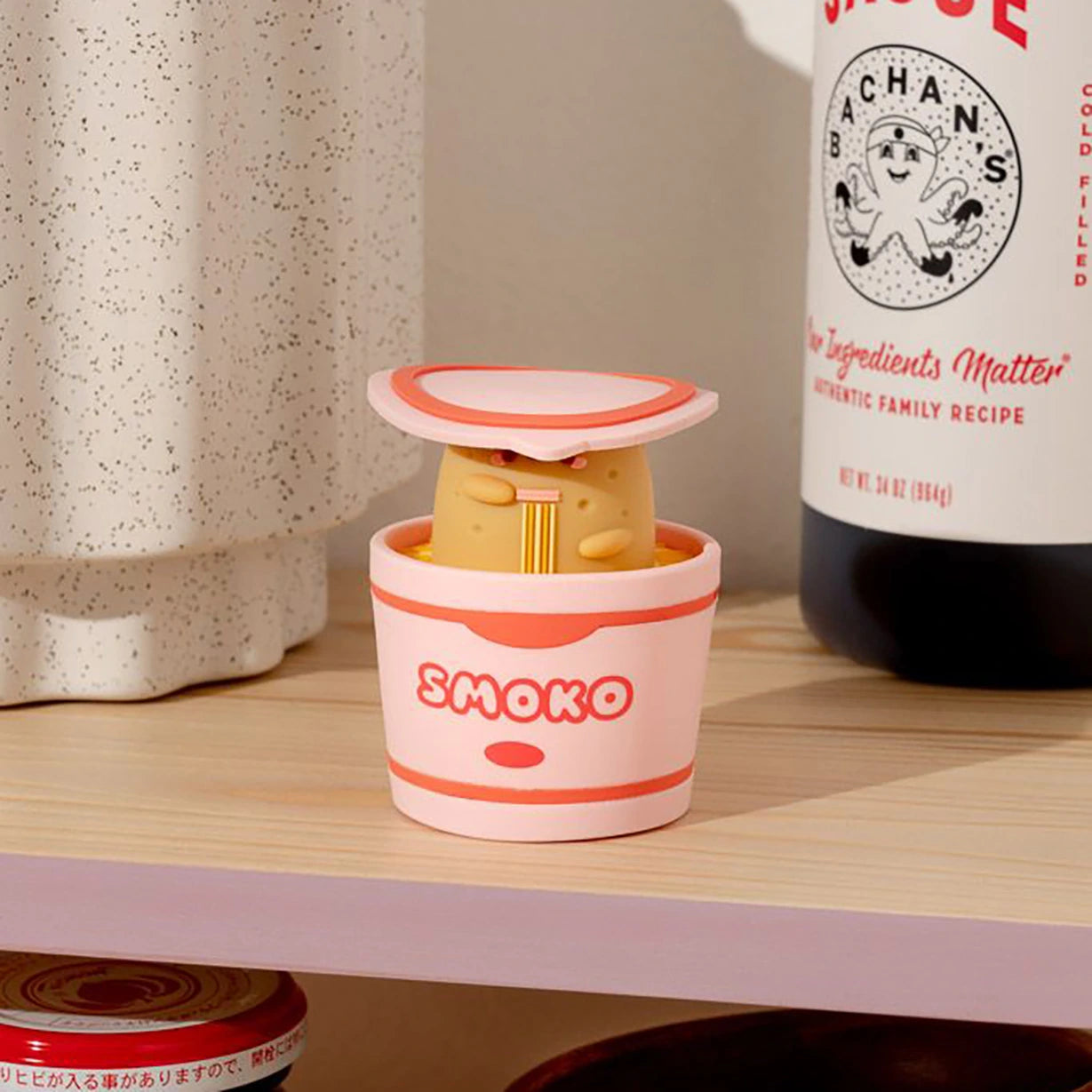 A charming figurine from the "Smoko Tayto Potato Ramen Blind Box" peeks out from a pink container on a wooden shelf, nestled next to a bottle and a textured ceramic piece, perfectly encapsulating the allure of vinyl figures.