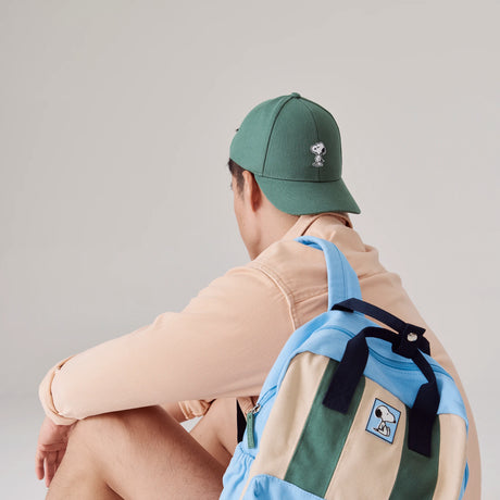 Green canvas baseball cap with Snoopy embroidered on front worn by seated person with colorful backpack