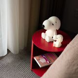 Two Snoopy Night Light lamps (11cm) sit on a red round side table by the couch. A Peanuts book lies on the lower shelf. The setting, with BPA-free silicone accessories, is near a window with sheer curtains.