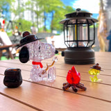 The Snoopy Camp 3D Crystal Puzzle features plastic models of a dog and bird in hats by a toy campfire and lantern on a wooden table, creating the perfect outdoor camping scene. An LED light casts a warm glow amid trees and buildings, enhancing this charming miniature setting.