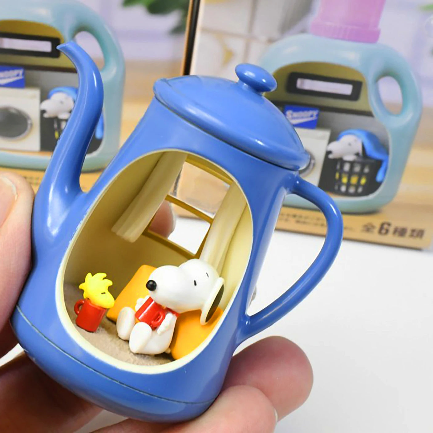 Small blue teapot figurine with Snoopy and Woodstock holding red mugs inside cozy miniature room setting