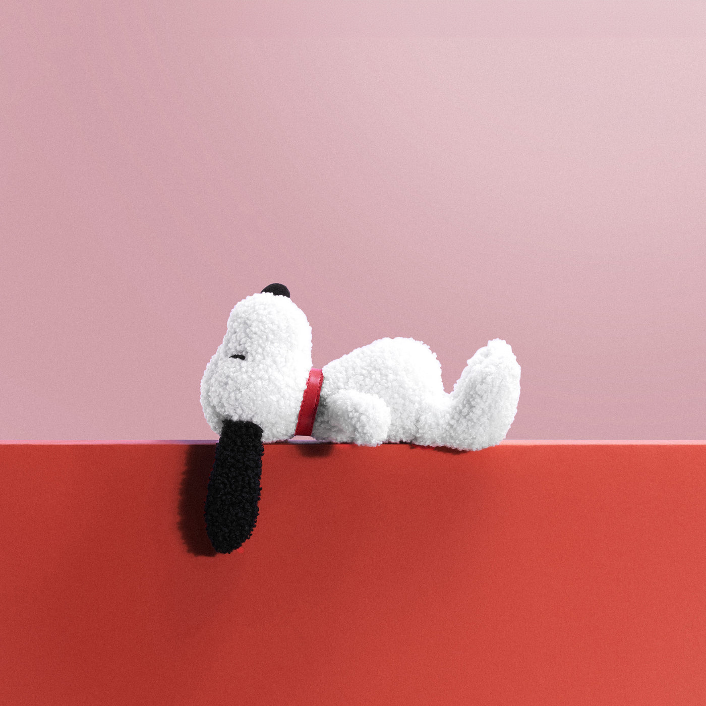 A 17cm Snoopy plush toy from the Snoopy Plush Doghouse Gift Set, featuring floppy ears and a red collar, rests elegantly on a crimson surface with a pink background, capturing the charm of Peanuts Magic.