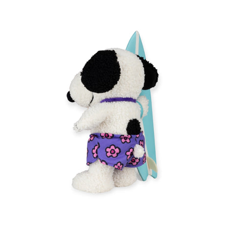 Rear view of Snoopy plush in purple floral board shorts holding blue surfboard standing on white background