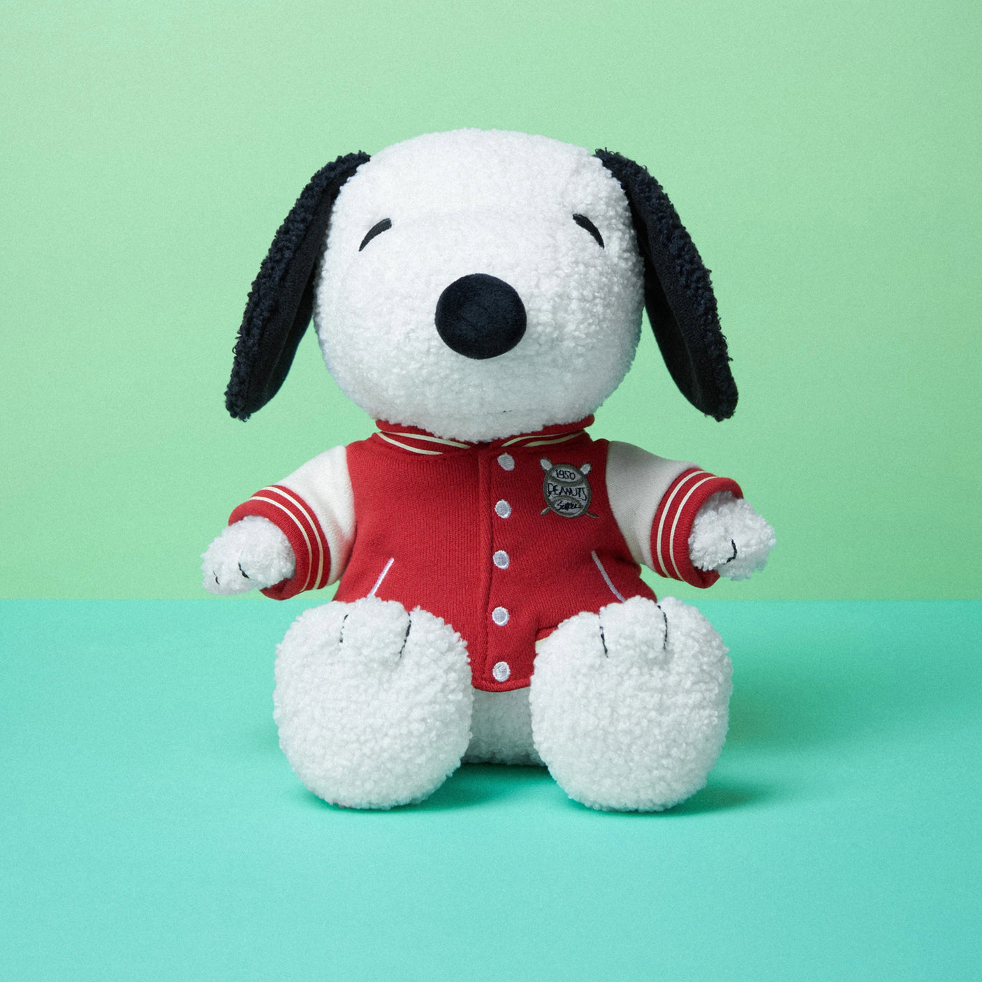 A Snoopy Plush Varsity Jacket 22cm - Limited Edition from the Snoopy brand is positioned upright against a two-tone green backdrop, proudly adorned in a red varsity jacket with white sleeves.