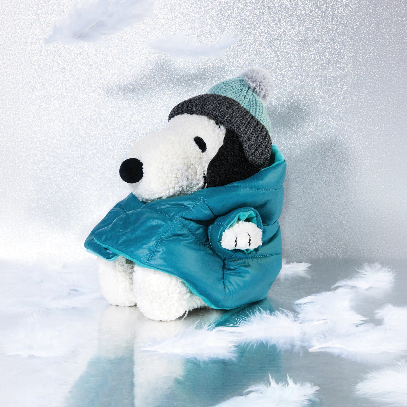 A limited edition 20cm Snoopy Plush from the Peanuts collection, wearing a teal puffer jacket and knit hat, rests amid white feathers on a shiny surface.