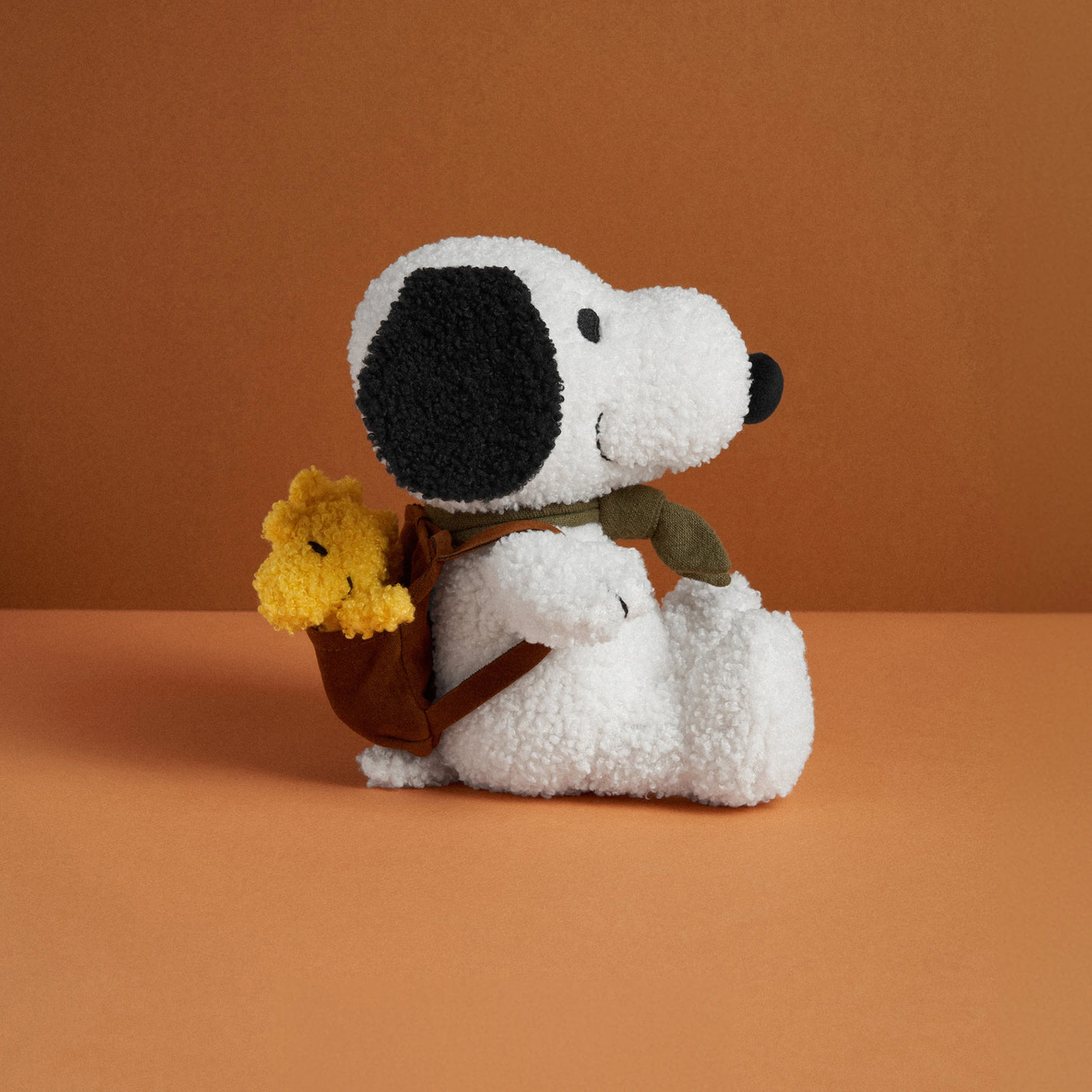 A Snoopy Plush Woodstock Backpack from the Snoopy brand, featuring black ears and nose, sits against an orange background. It's adorned with a green scarf and carries a small brown bag with a yellow flower.