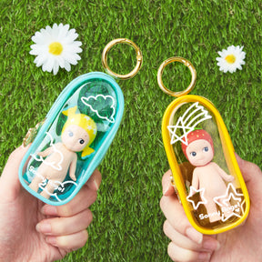 Two hands holding Sonny Angel My Bestie Cases in yellow and turquoise with star and cloud designs on grass background