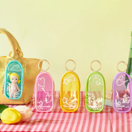 The Sonny Angel My Bestie Case - Purple by Sonny Angel is shown on a red checkered cloth with its white ribbon design, one attached to a beige bag holding a small doll—ideal as a mini figure carry case.