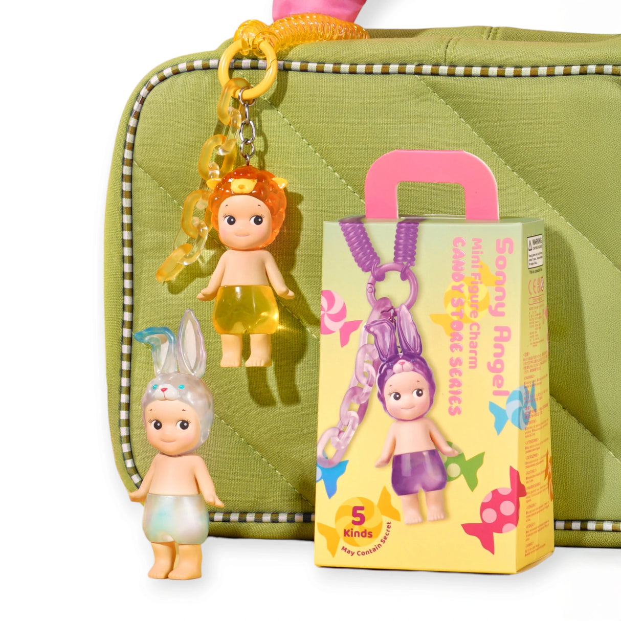 Two small figurines from the Sonny Angel Candy Store Series, one with a lion headpiece and the other with bunny ears, are next to a decorative Blind Box by Sonny Angel attached to a green quilted bag with a yellow chain.