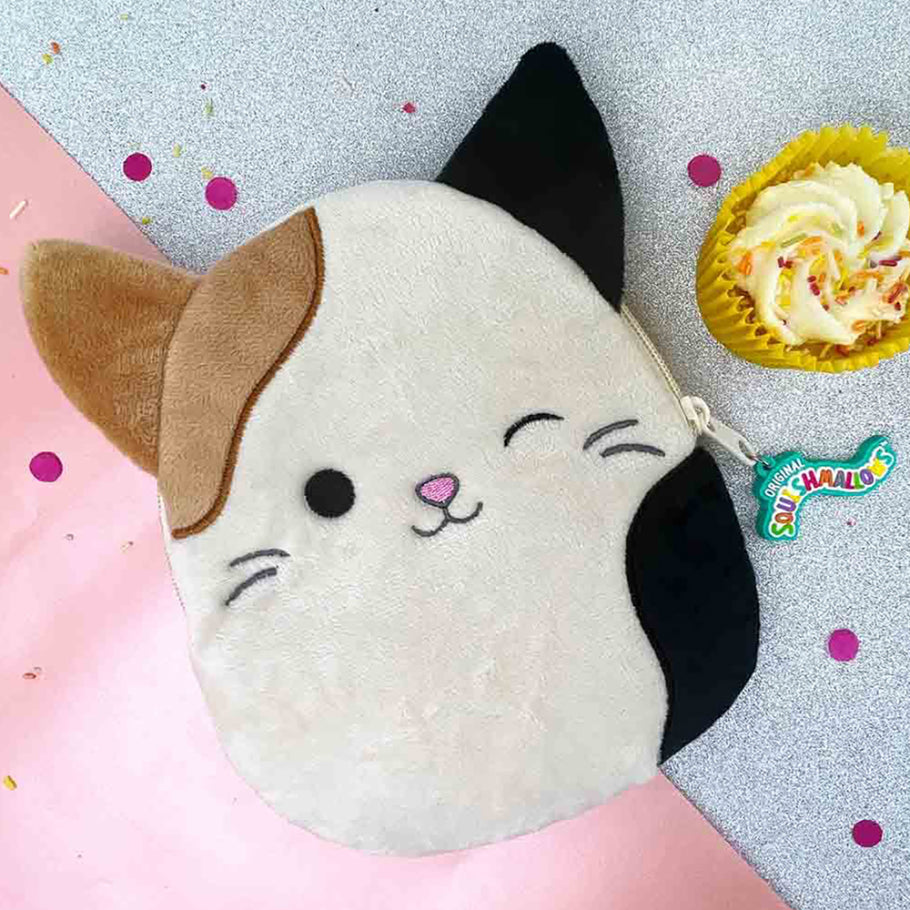 The Squishmallows Calico Cat Plush Case, featuring embroidered details, sits on a multicolored surface. It is accompanied by a yellow cupcake and candies labeled "Sour Smog Balls," making it an ideal playful pencil case or plush holder for your essentials.