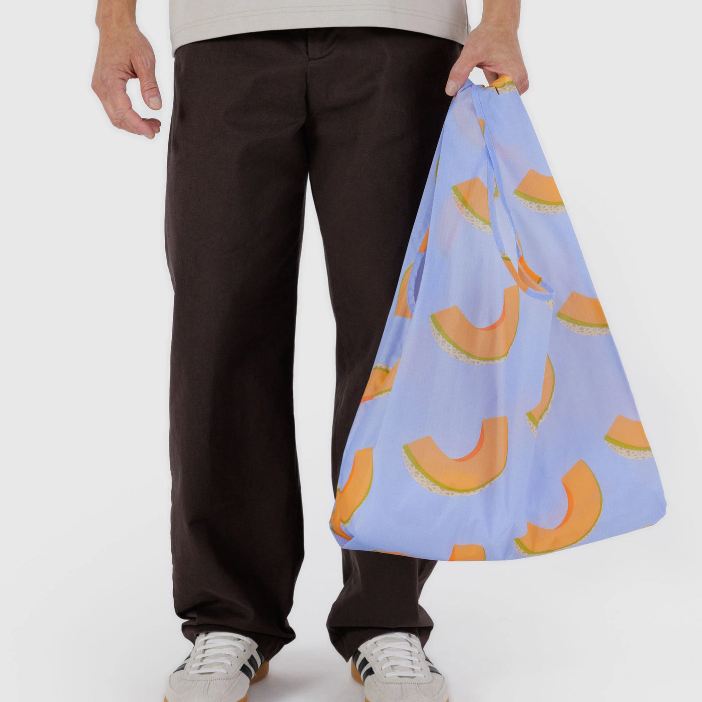 A person carries a Baggu Standard Bag, the Cantaloupe Blue version, while dressed in a light shirt, dark pants, and white sneakers. Made from recycled ripstop nylon, this bag is stylish and eco-friendly.