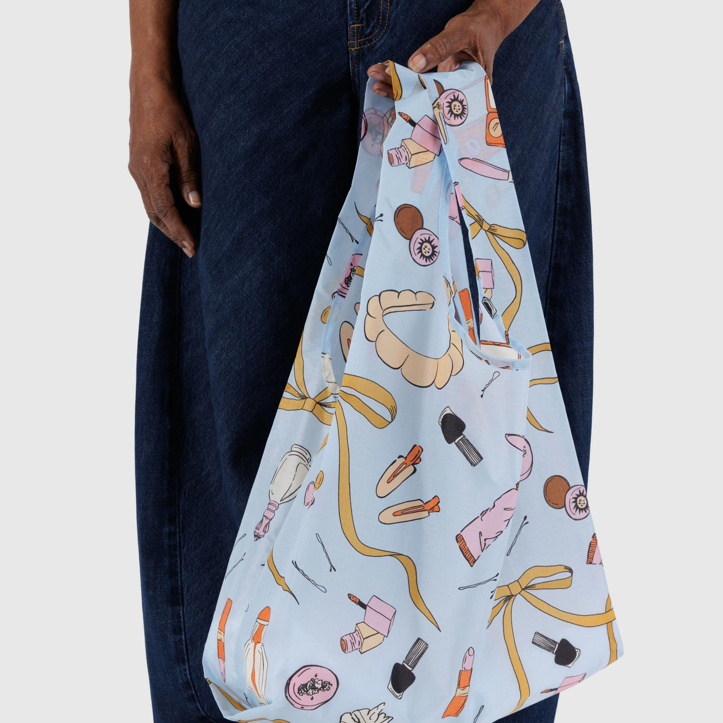 A person carries the Baggu Standard Bag - Get Ready With Me, designed in light blue recycled ripstop nylon with playful cosmetics and accessories prints, paired with dark pants.