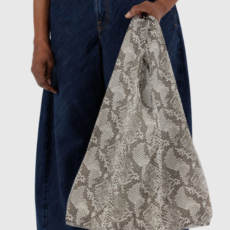 A person carries the Standard Baggu Bag - Snakeskin from Baggu, crafted with recycled ripstop nylon, while wearing dark blue denim pants against a light gray background.