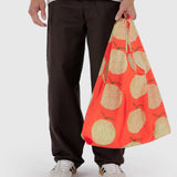 A person holds a Standard Baggu Bag - Yubari by Baggu, featuring apple prints on recycled ripstop nylon, while wearing brown pants and white sneakers.