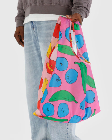 An individual is showcasing the Baggu Standard Reusable Bag - Apples & Bananas by Baggu, featuring vibrant fruit patterns of bananas and apples, set against a plain background. This eco-chic tote bag merges stylish flair with sustainable design, ideal for the fashionista who values environmental responsibility.