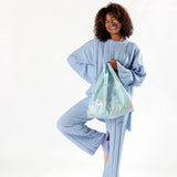 A person in a blue outfit stands on one leg, smiling, while holding a Baggu Standard Bag featuring Cinnamoroll.