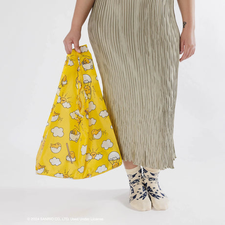 A person in a pleated skirt is holding a vibrant yellow Baggu Standard Bag - Gudetama featuring an adorable Gudetama egg design, paired with patterned socks. Made by Baggu, the bag is crafted from recycled polyester, bringing an eco-friendly element to the quirky ensemble.