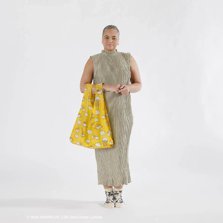 A person in a long dress is holding a Baggu Standard Bag - Gudetama, featuring a yellow pattern, while standing against a plain white background.